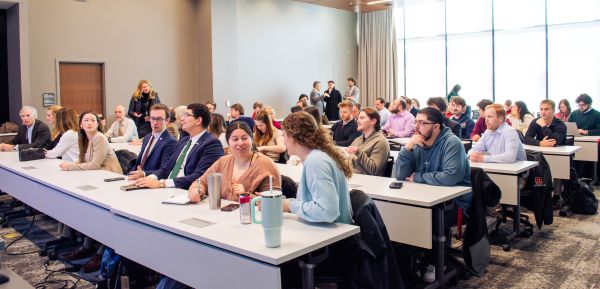 Approximately sixty people sit at desks in long rows. Some engage in conversation