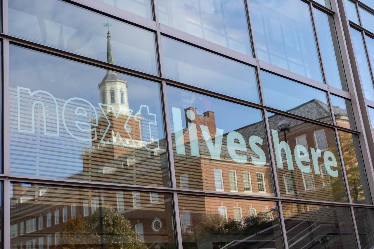 Next Lives Here written on a building with Arts and Sciences Hall reflection on it